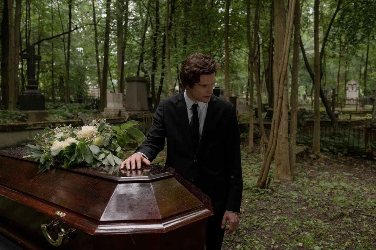 story-01 A man in a black suit mourns at a funeral in a lush green cemetery, leaning on the coffin.